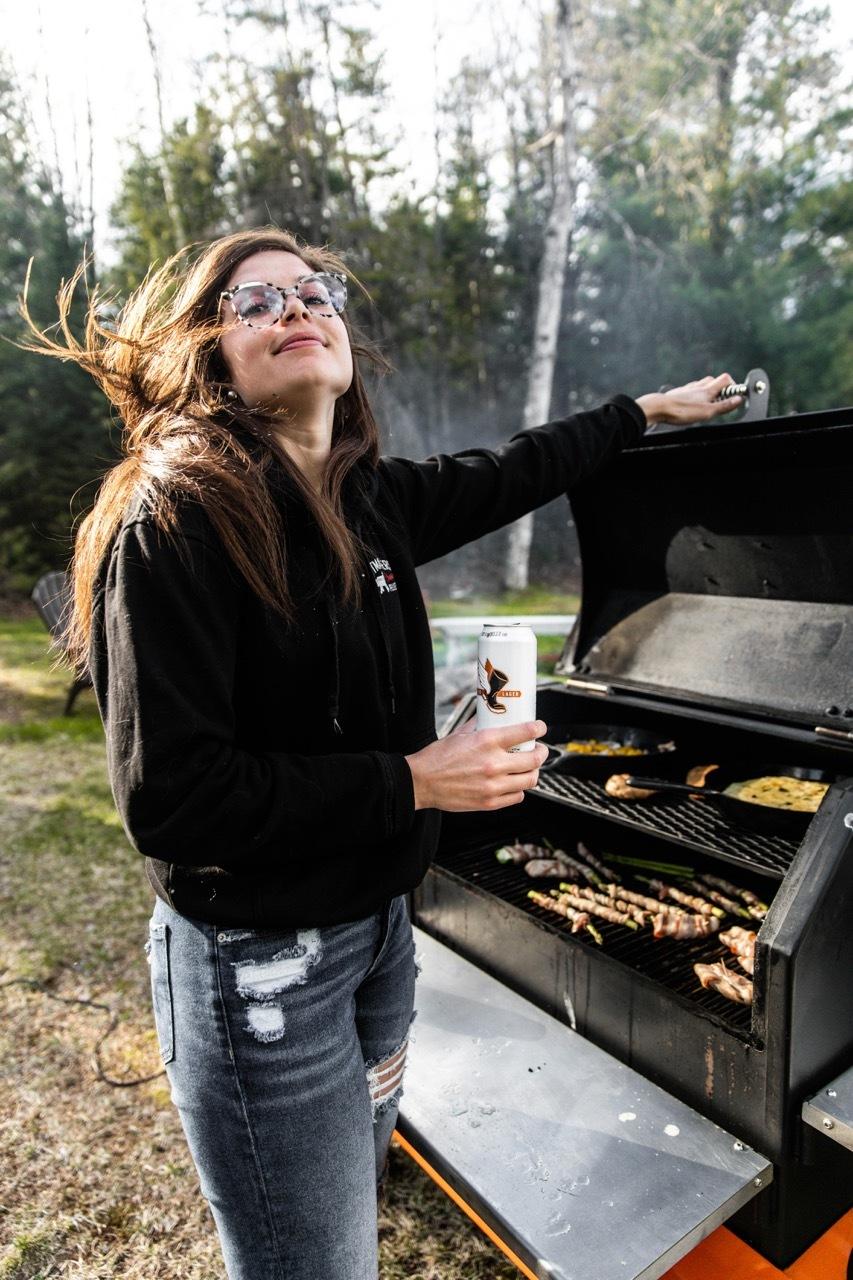 Woman holding a drink beside an open outdoor grill.