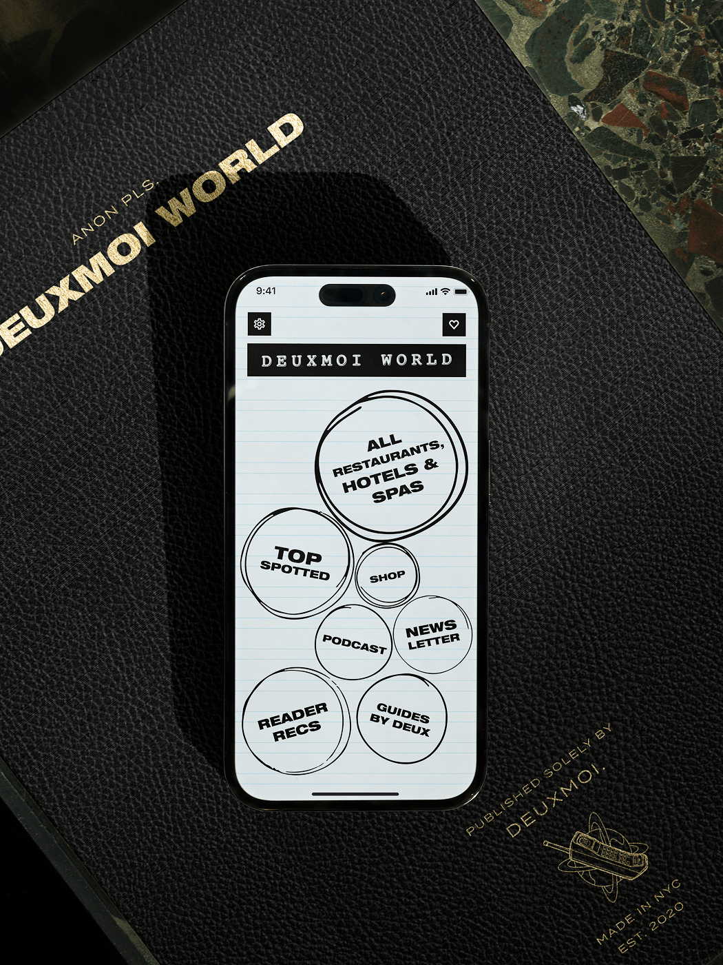 Phone showing Deuxmoi World app menu on a black leather book