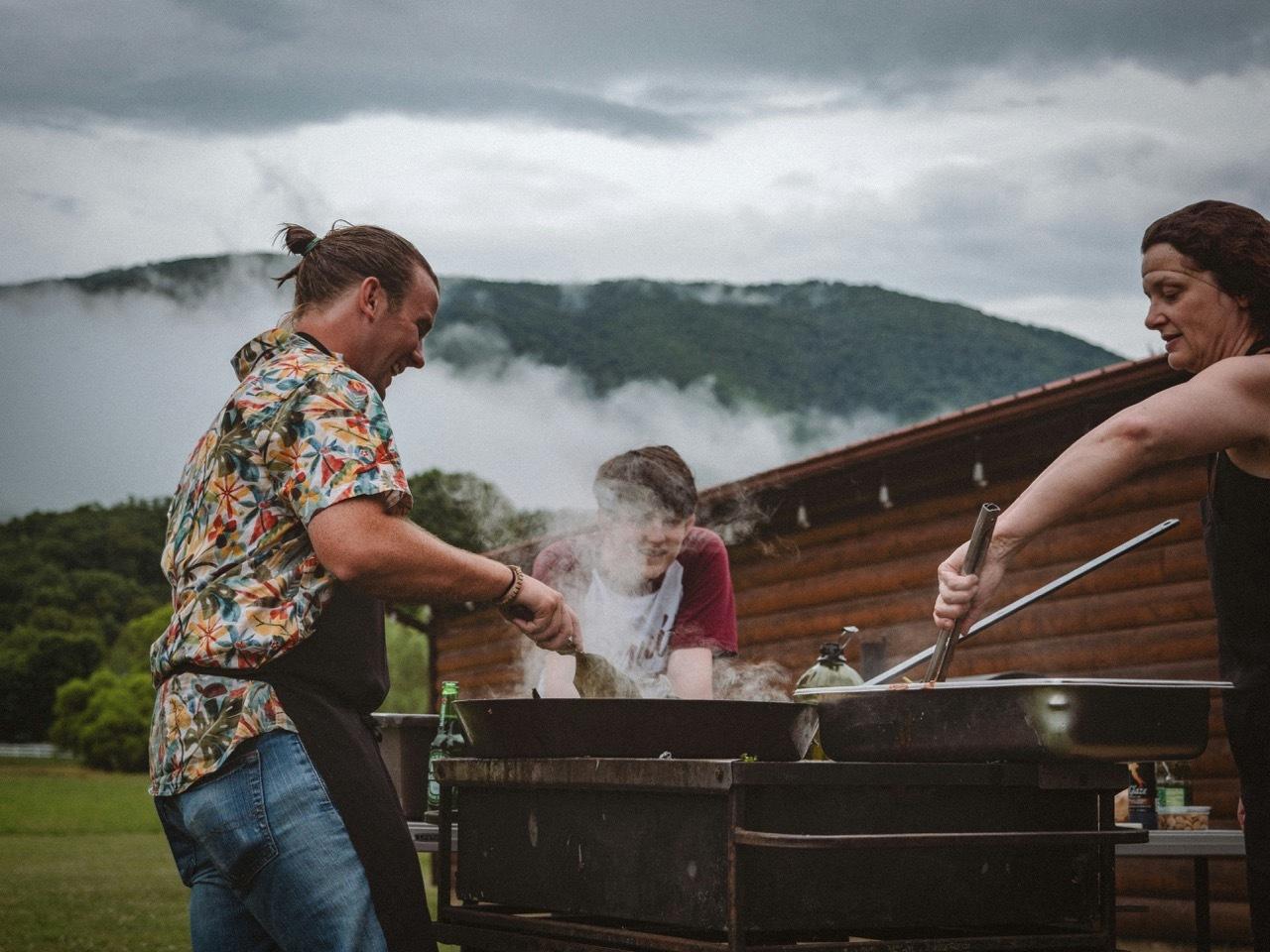 People cooking at an outdoor grill with misty mountains behind them.