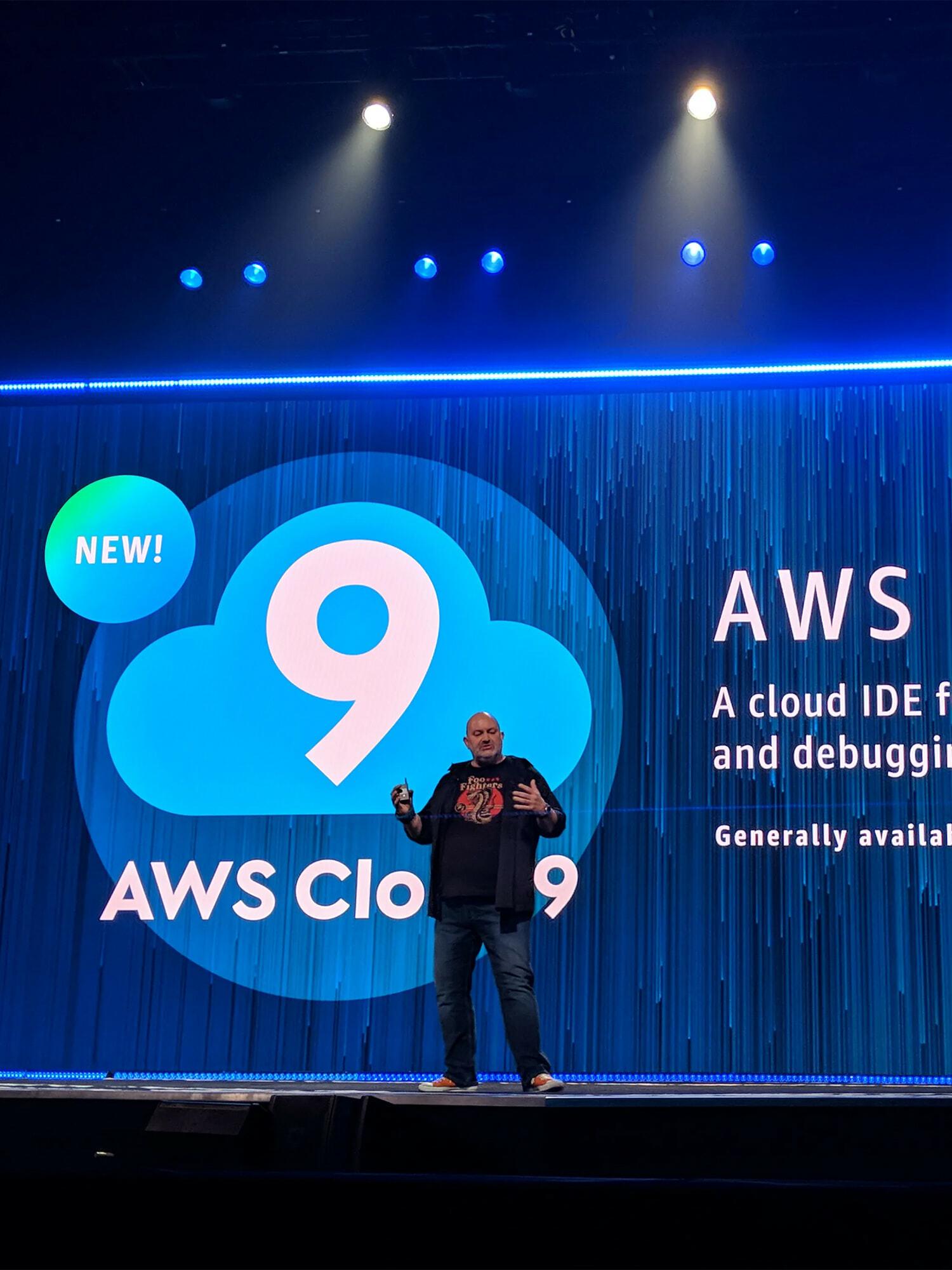 Presenter on stage before a large AWS Cloud9 slide with blue lighting and spotlights.