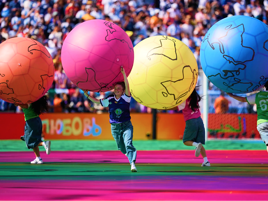 Performers carry oversized colorful soccer balls across a stadium field.