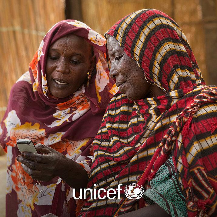 Two women in patterned headscarves looking at a phone with UNICEF logo overlay.