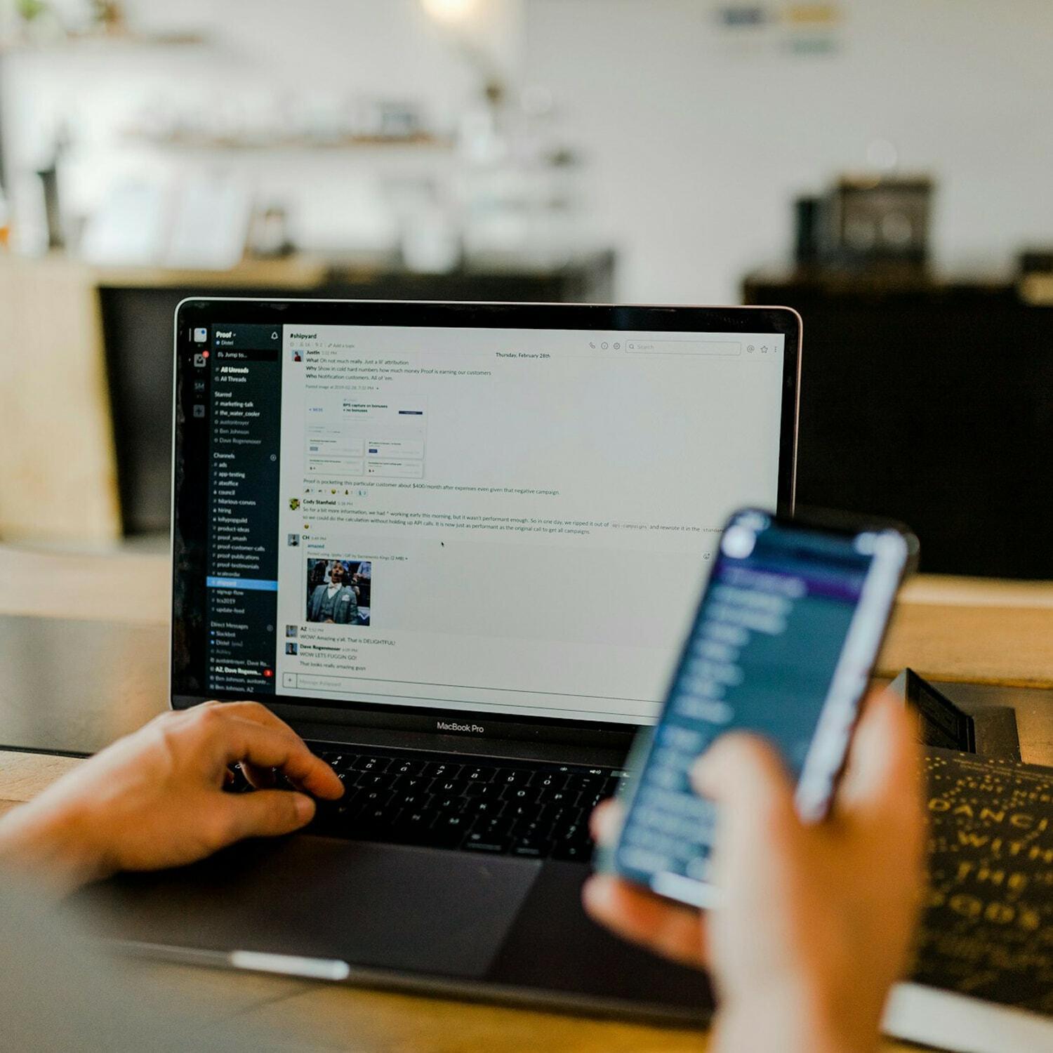 Hands using Slack on a MacBook Pro and a smartphone at a cafe table.