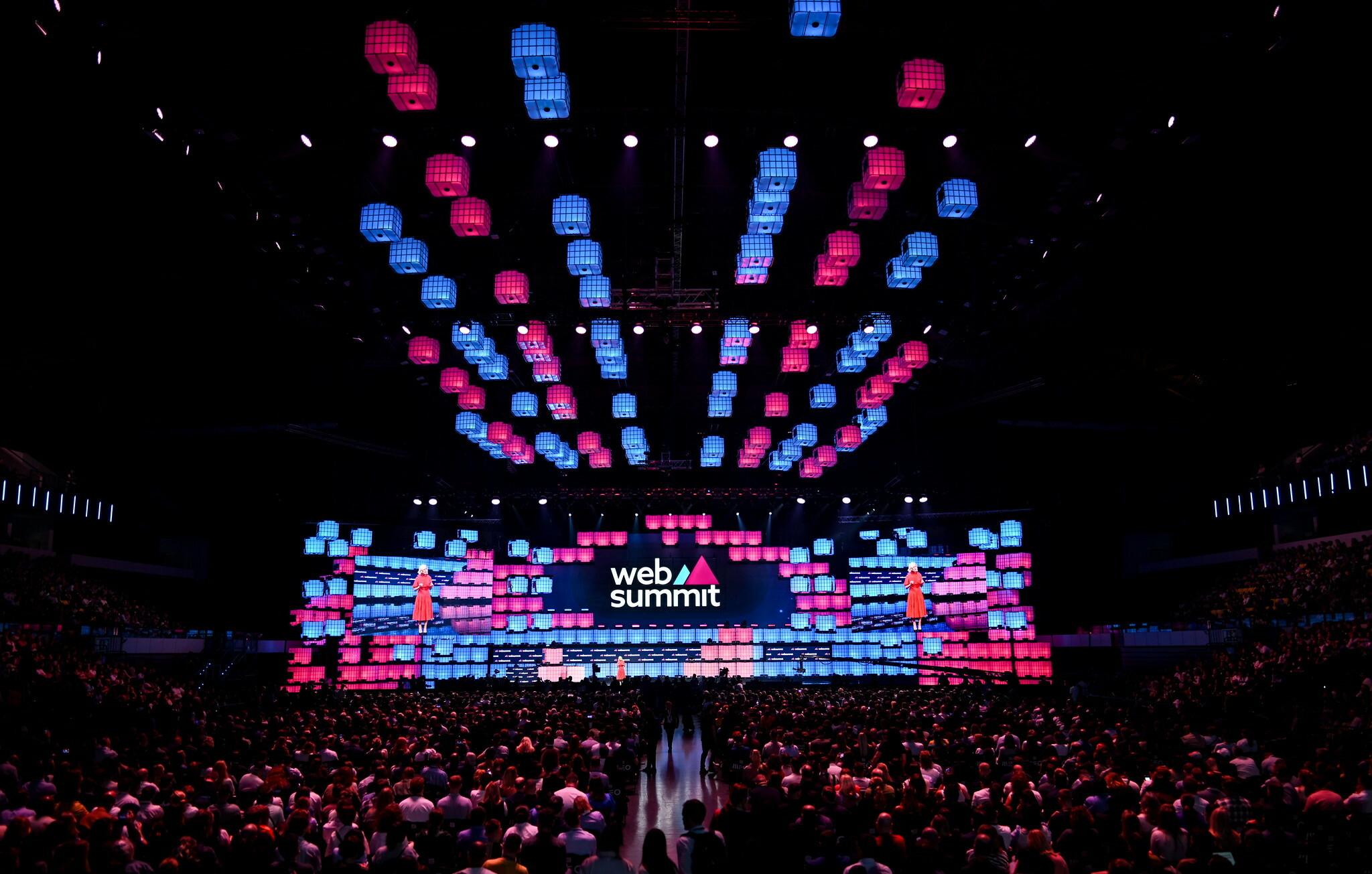 Web Summit main stage lit in pink and blue LED panels above a packed arena